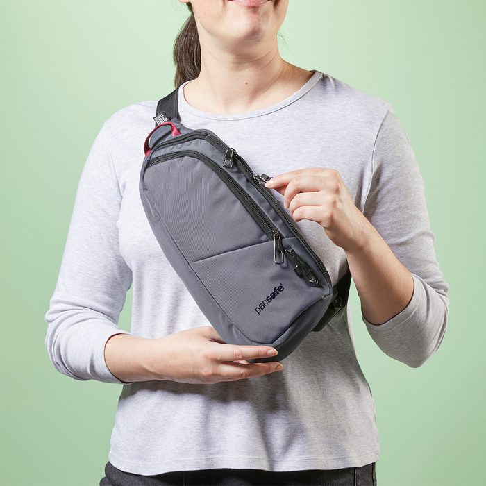 A person wears a gray sling bag over their shoulder, adjusting it against a light green background.