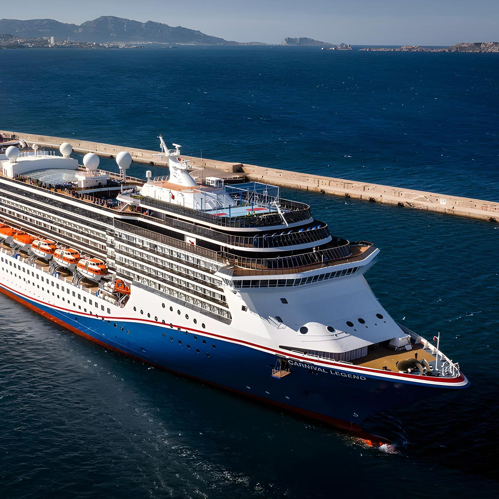 Cruise ship navigating near a coastal dock, on clear blue ocean waters, with distant mountainous landscape in the background. Text reads "Carnival Legend."