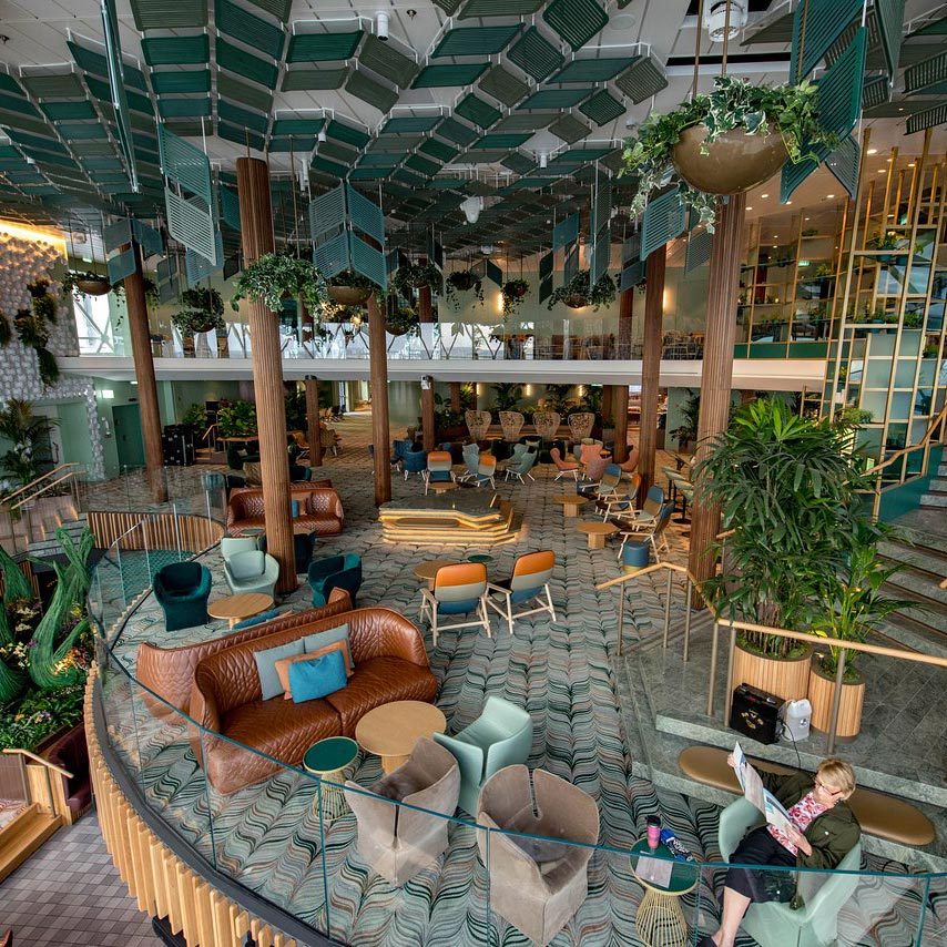 Furniture fills a modern, spacious lounge with plants, patterned flooring, and a person reading. Geometric ceiling decor adds an artistic touch.