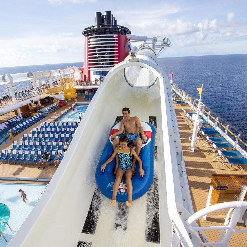 Two people ride a waterslide raft aboard a cruise ship, surrounded by deck chairs and pools, with a vast ocean background.