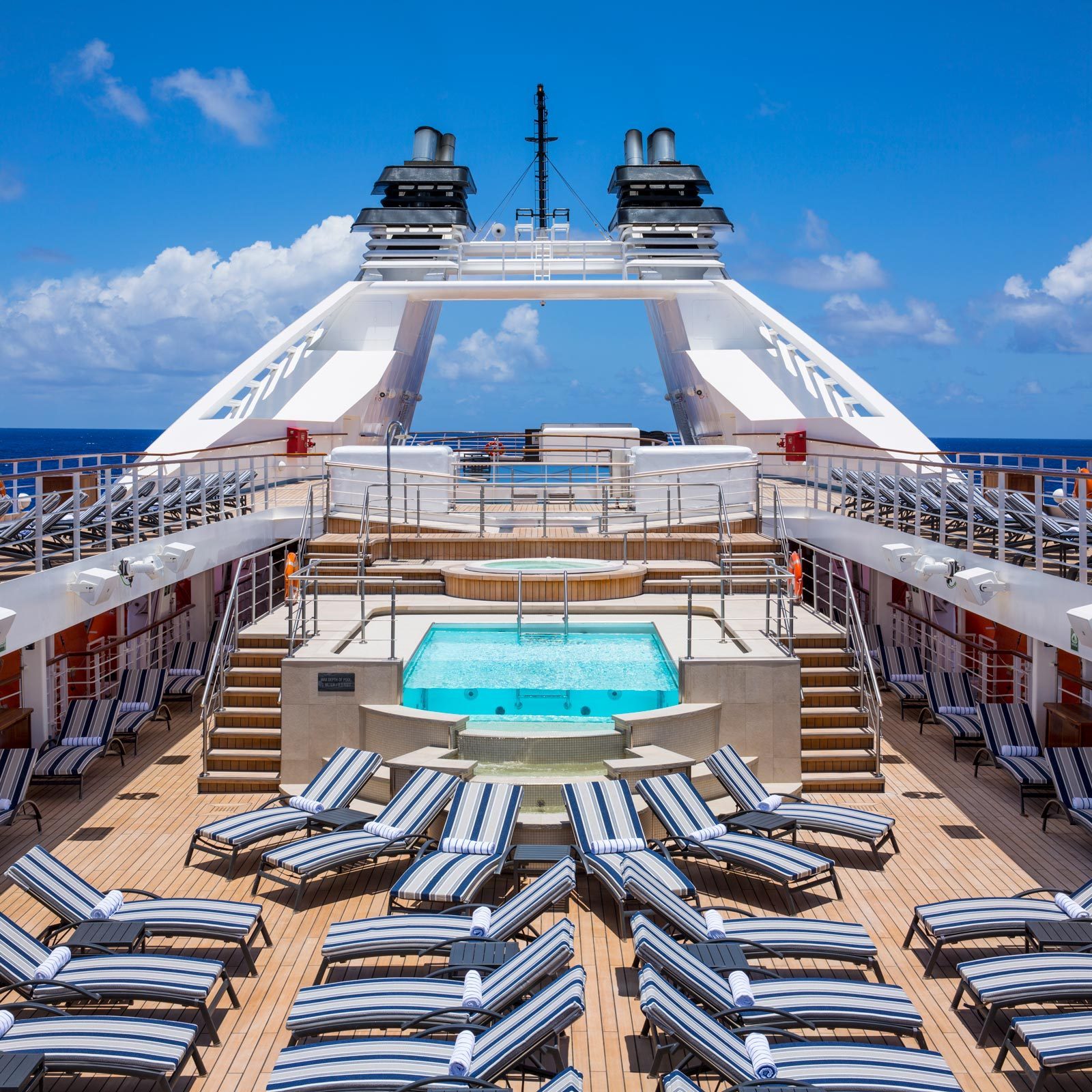 Pool sits amidst striped loungers on cruise ship deck, clear blue sky above, and ocean in the background.