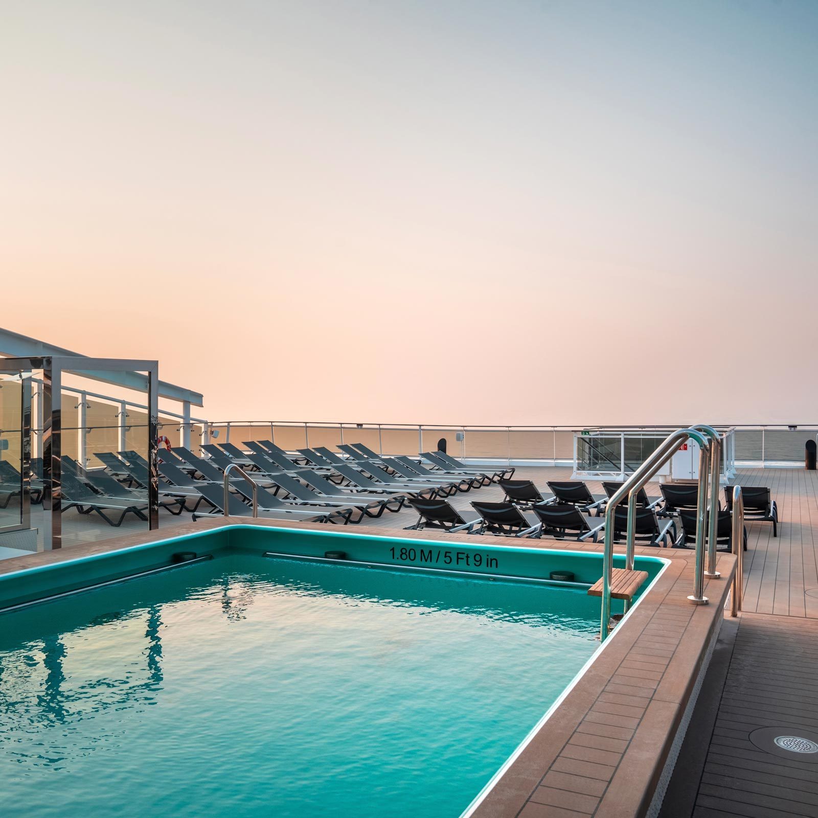 Pool reflects soft pastel sky; surrounded by empty lounge chairs on a cruise ship deck. Depth marking reads "1.80 M / 5 Ft 9 in."