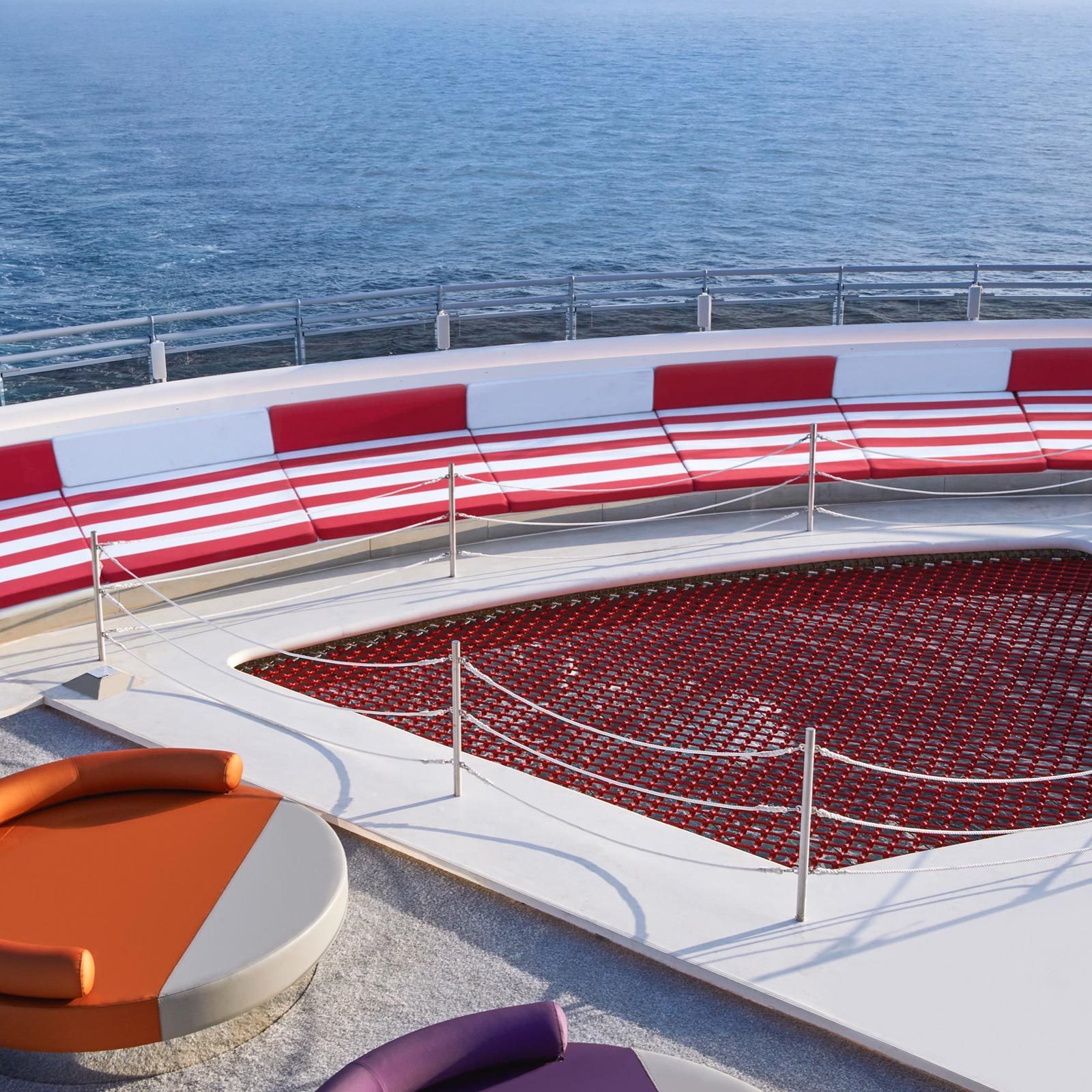 Netting covers a red trampoline surrounded by striped white and red seating on a ship deck, with the ocean in the background.