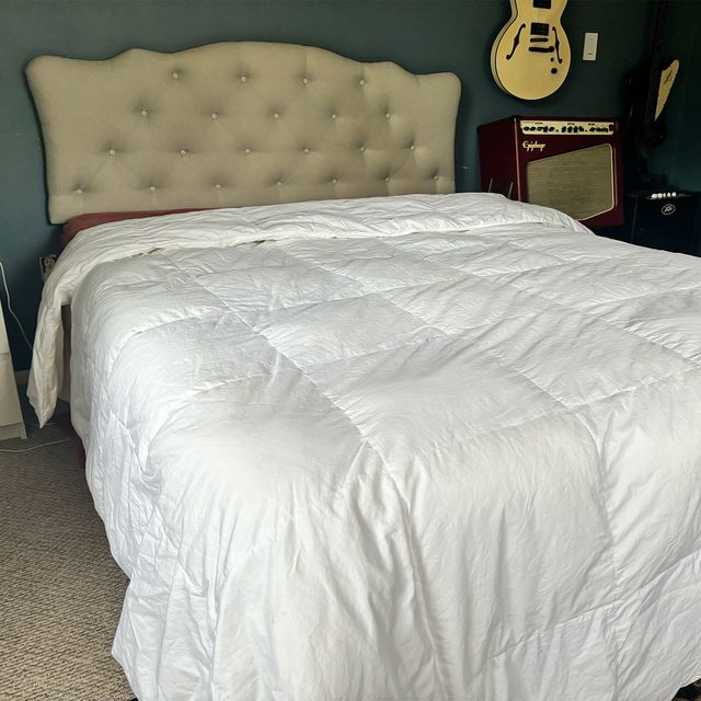 Bed with white comforter sits in a room; tan headboard behind, guitar and amplifier on the side.