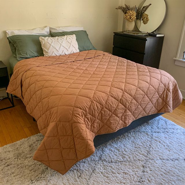 A bed covered with a brown quilt is in a cozy bedroom next to a dresser with a mirror and dried flowers.