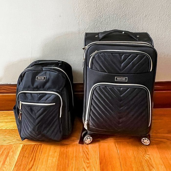Backpack and rolling suitcase standing together on wooden floor, against a light-colored wall. Both are black with quilted patterns and silver zippers.
