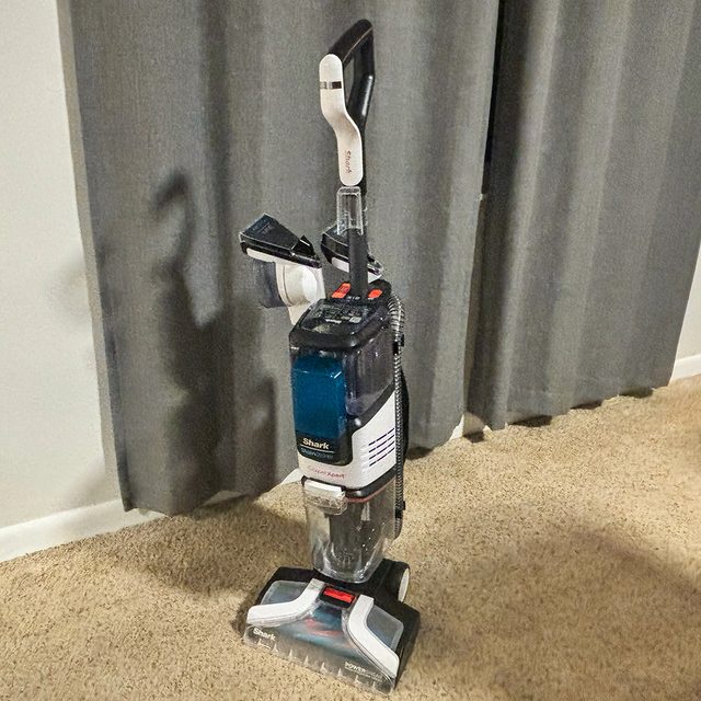 Vacuum cleaner stands upright on carpeted floor, positioned near a grey curtain.