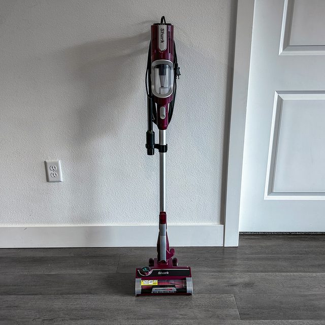 A vacuum cleaner stands upright against a wall on a wooden floor, with cord coiled neatly.