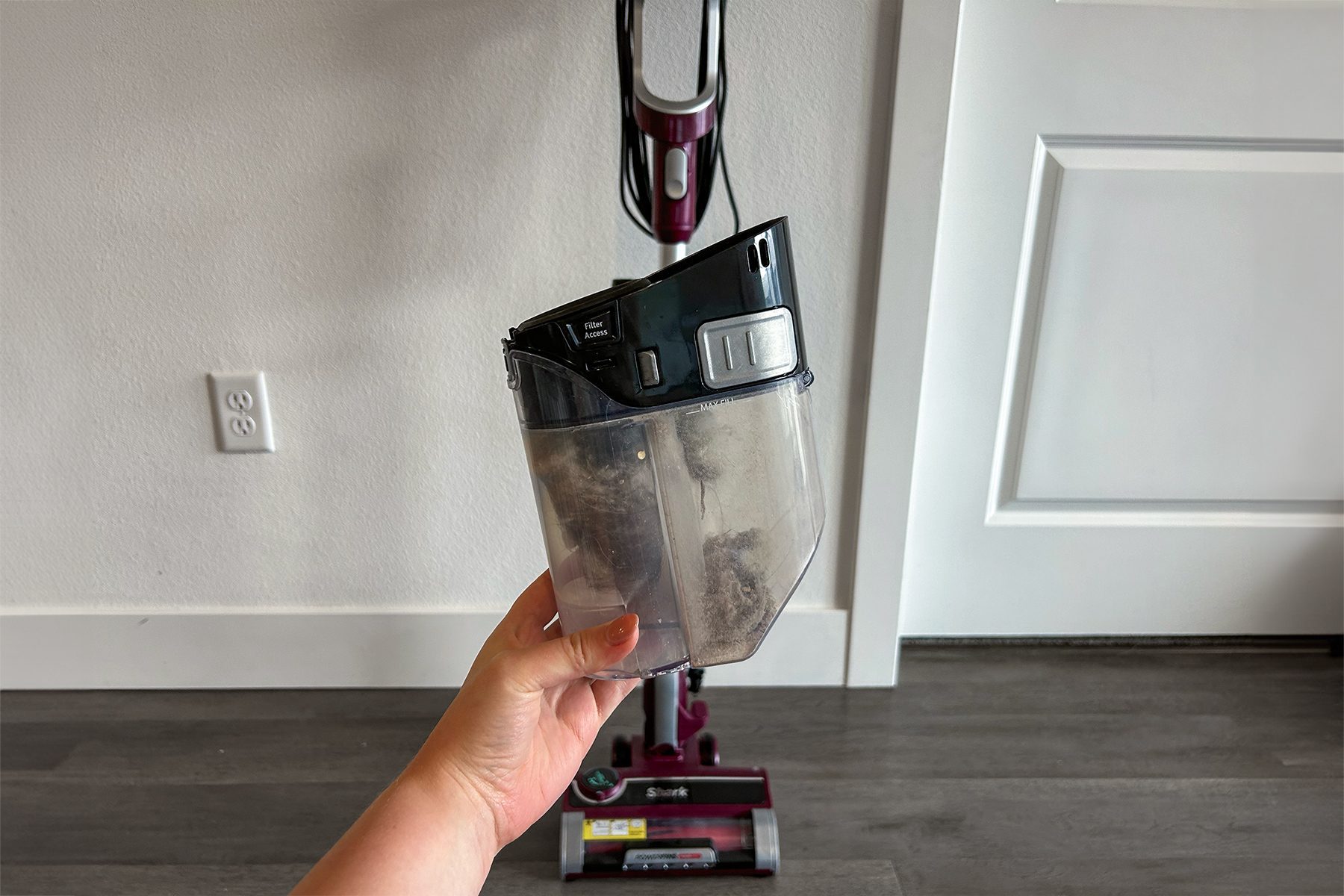 Hand holds a vacuum dust container; vacuum stands on a wooden floor near a white wall and door. "Shark" is visible on the vacuum base.