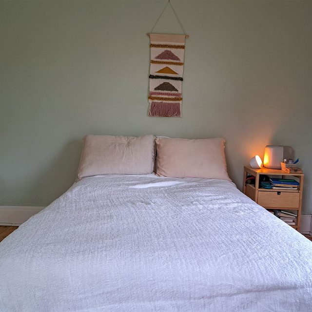 Bed with white covers is neatly arranged, beside a small nightstand with a glowing lamp. A decorative wall hanging adorns the light-colored wall.