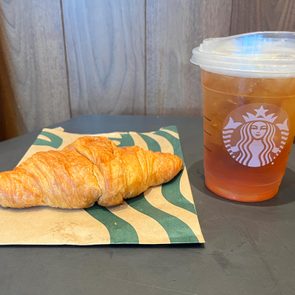Croissant rests on patterned paper; iced drink in clear Starbucks cup nearby; wood-paneled background.