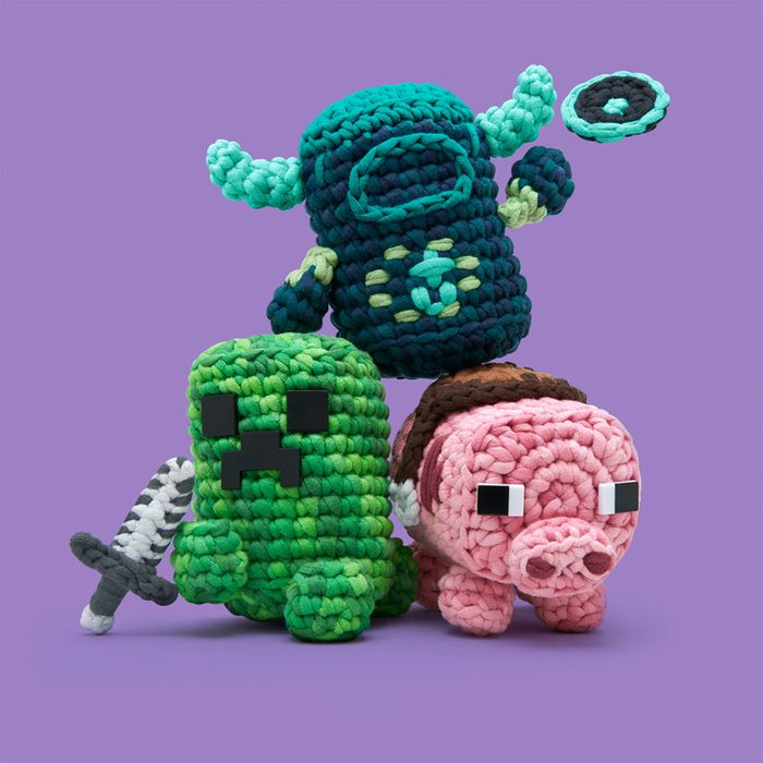 Three crocheted toy figures, resembling game characters, stand together on a purple background; one holds a sword.