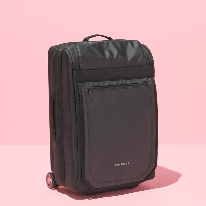 A black suitcase stands, displaying its front pocket on a pink background. Text reads 