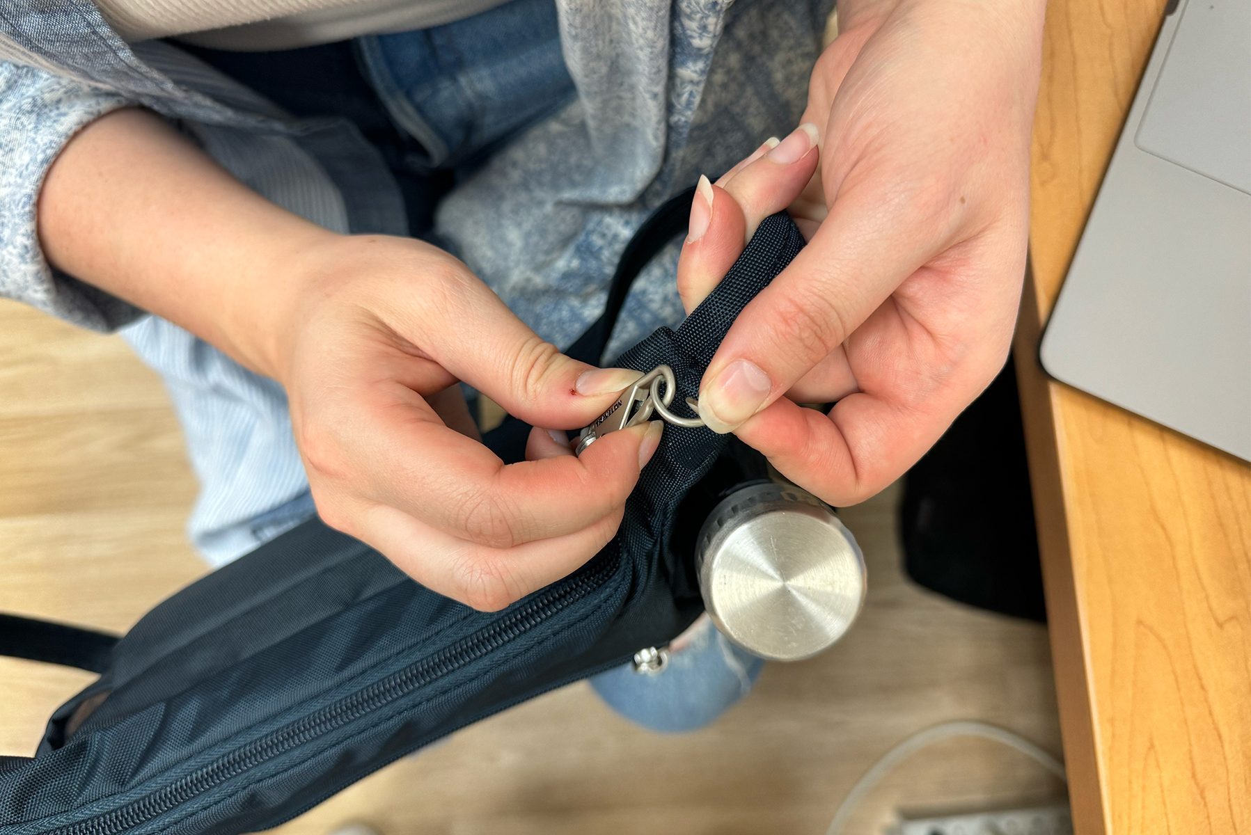 Hands attaching a keyring to a backpack loop, next to a stainless steel bottle, with a wooden desk and laptop nearby.