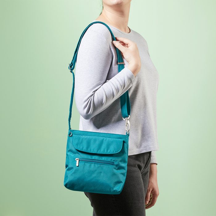 A woman carries a teal shoulder bag against a light green background, wearing a gray long-sleeve shirt and dark pants.