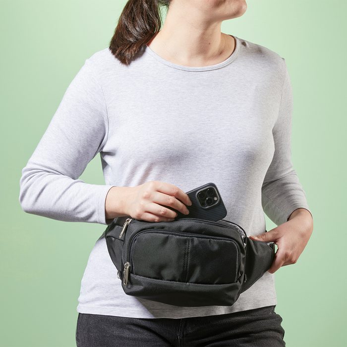 A person places a smartphone into a black waist bag, standing against a light green background.