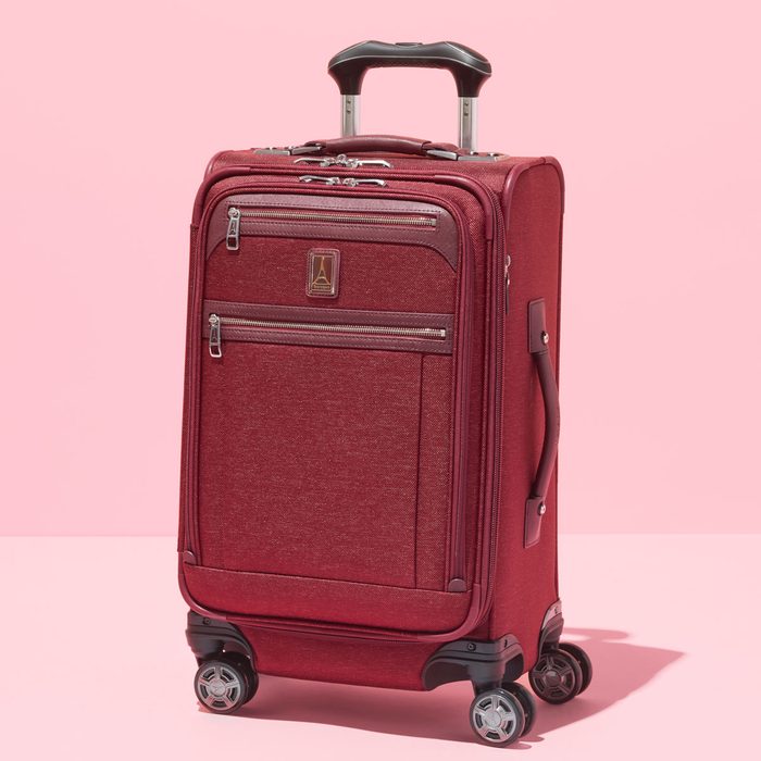 A red suitcase stands upright with extended handle on four wheels against a pink background.