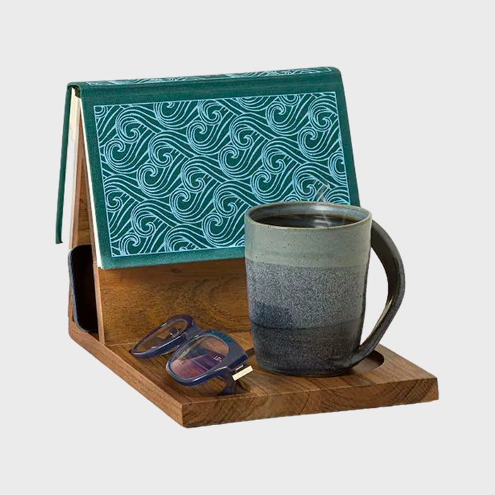 A mug rests on a wooden tray next to eyeglasses; behind, a book with a turquoise wave pattern stands open like a tent.