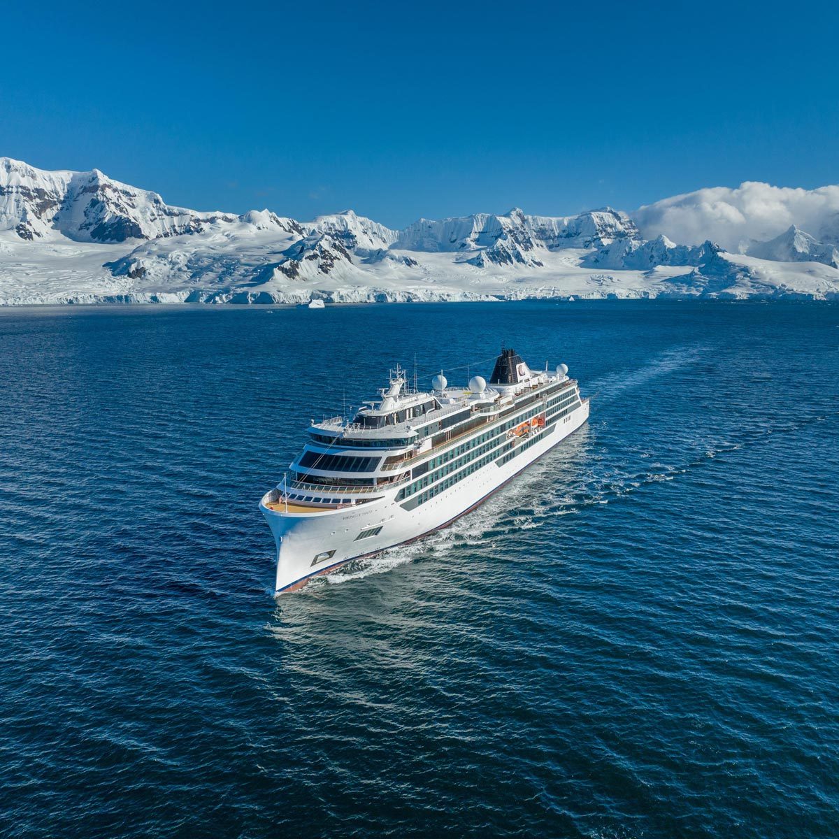 A cruise ship sails through calm blue waters, surrounded by snow-covered mountains under a clear blue sky.