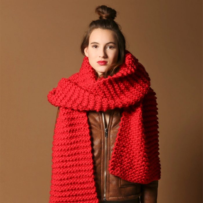 A person wears a large, chunky red scarf over a brown leather jacket against a plain brown background.