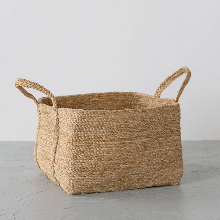 Woven basket sits on textured surface, with looped handles, against a plain white background.