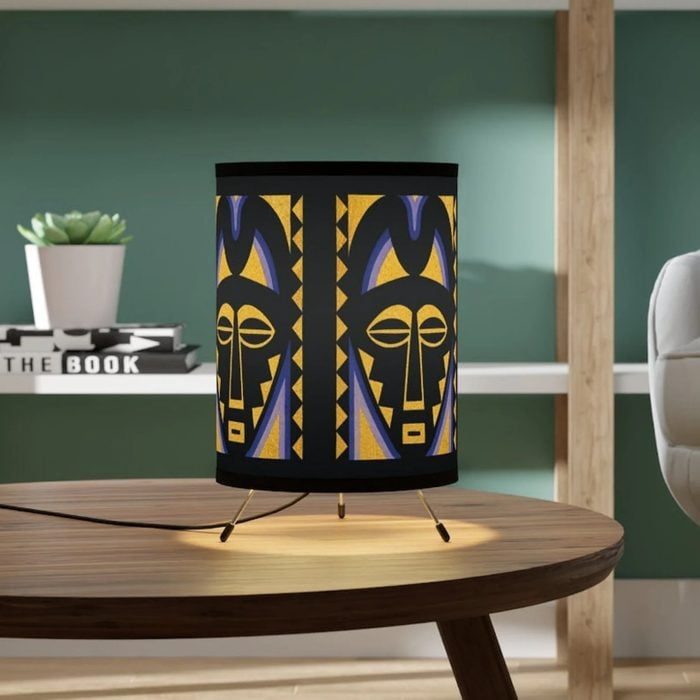 Lamp with tribal face design rests on a table; background includes a plant, books labeled 