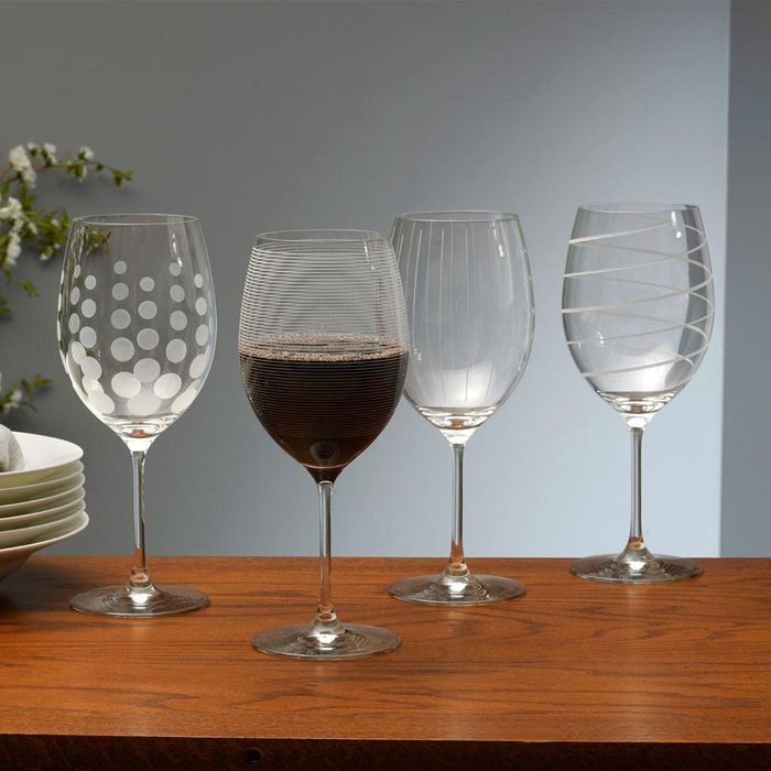 Four wine glasses with distinct patterns stand on a wooden table; one contains dark liquid. Soft lighting highlights the designs against a plain background.