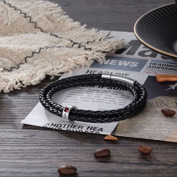 A black braided bracelet lies on a textured newspaper, surrounded by coffee beans and a fringed cloth on a wooden surface.