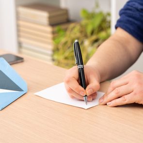A hand writes on paper with a pen. Nearby, an open blue envelope and a smartphone rest on a wooden table beside books and plants.