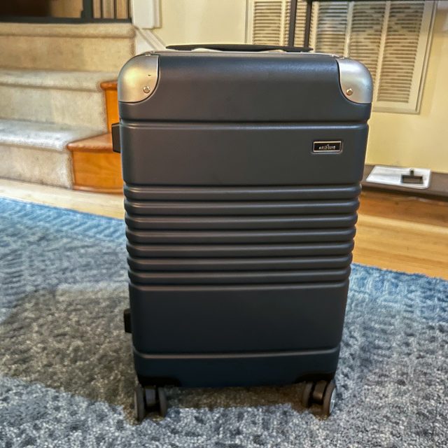 A black suitcase stands upright on a blue rug, with stairs and a vent in the background. Features silver corners and a nameplate reading 