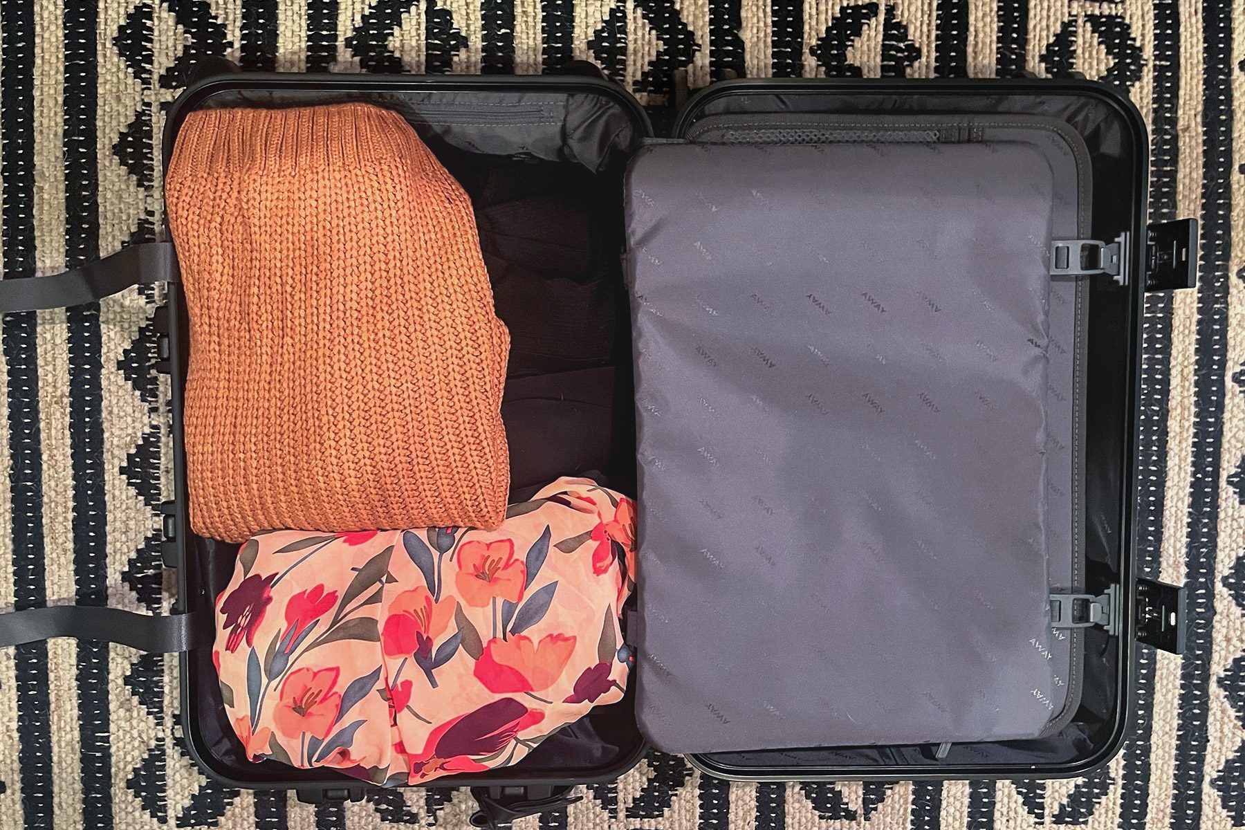 A suitcase holds folded clothes: an orange sweater and floral-patterned fabric, lying on a patterned rug.