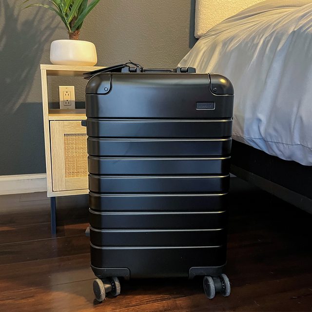 Black suitcase stands beside a nightstand within a bedroom featuring wooden floors and gray walls, showing a potted plant and white bed linens nearby.