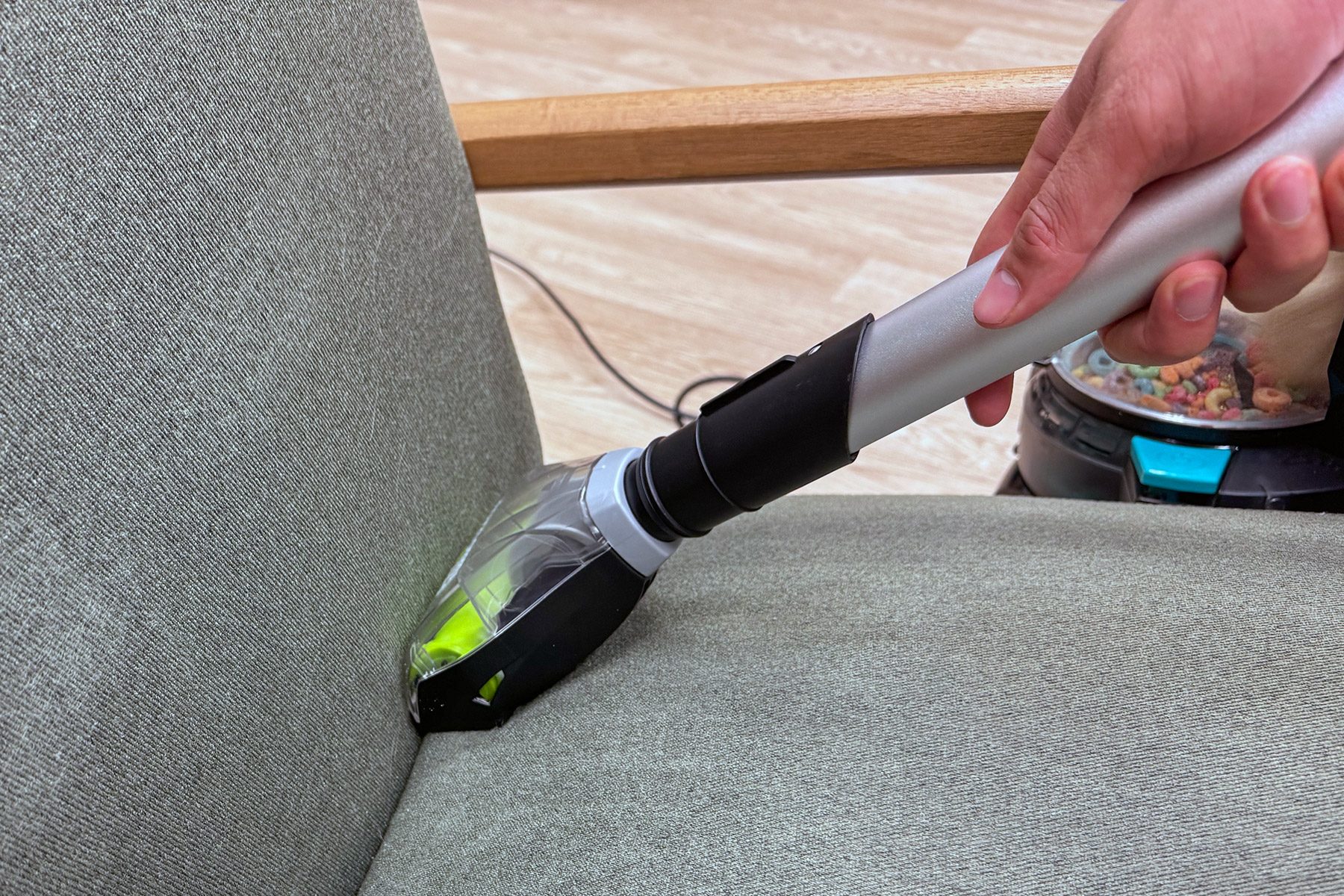 Vacuum head cleaning a gray upholstered chair, held by a hand, in a room with wooden flooring.