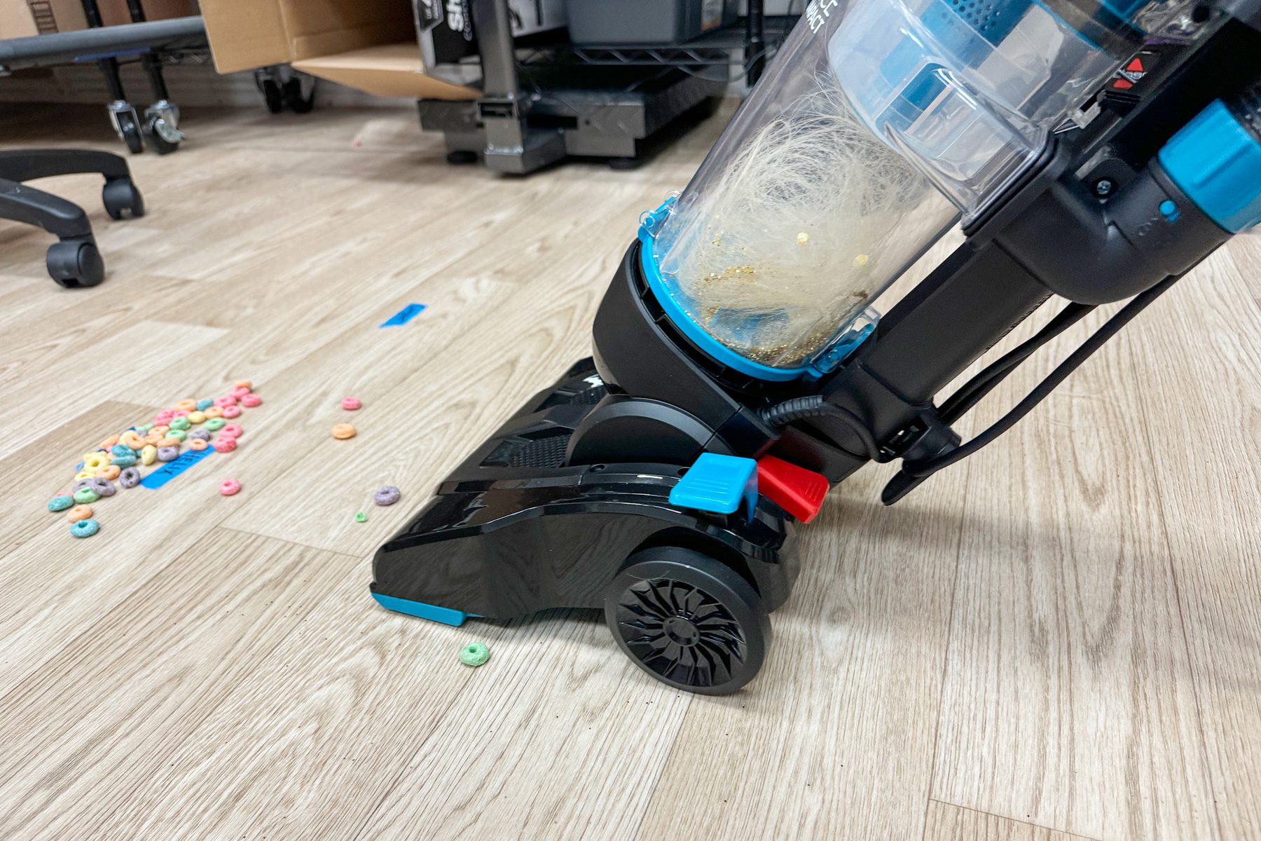 Vacuum cleaner collects cereal pieces on a wooden floor, with scattered fruit loops nearby in a cluttered room.