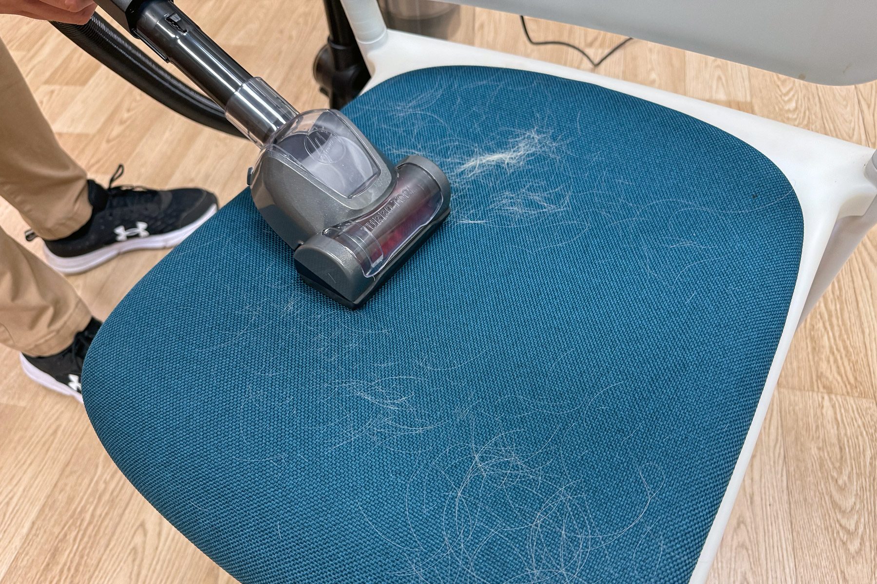 Vacuum removes pet hair from blue fabric chair, with wood flooring and person wearing sneakers visible nearby.