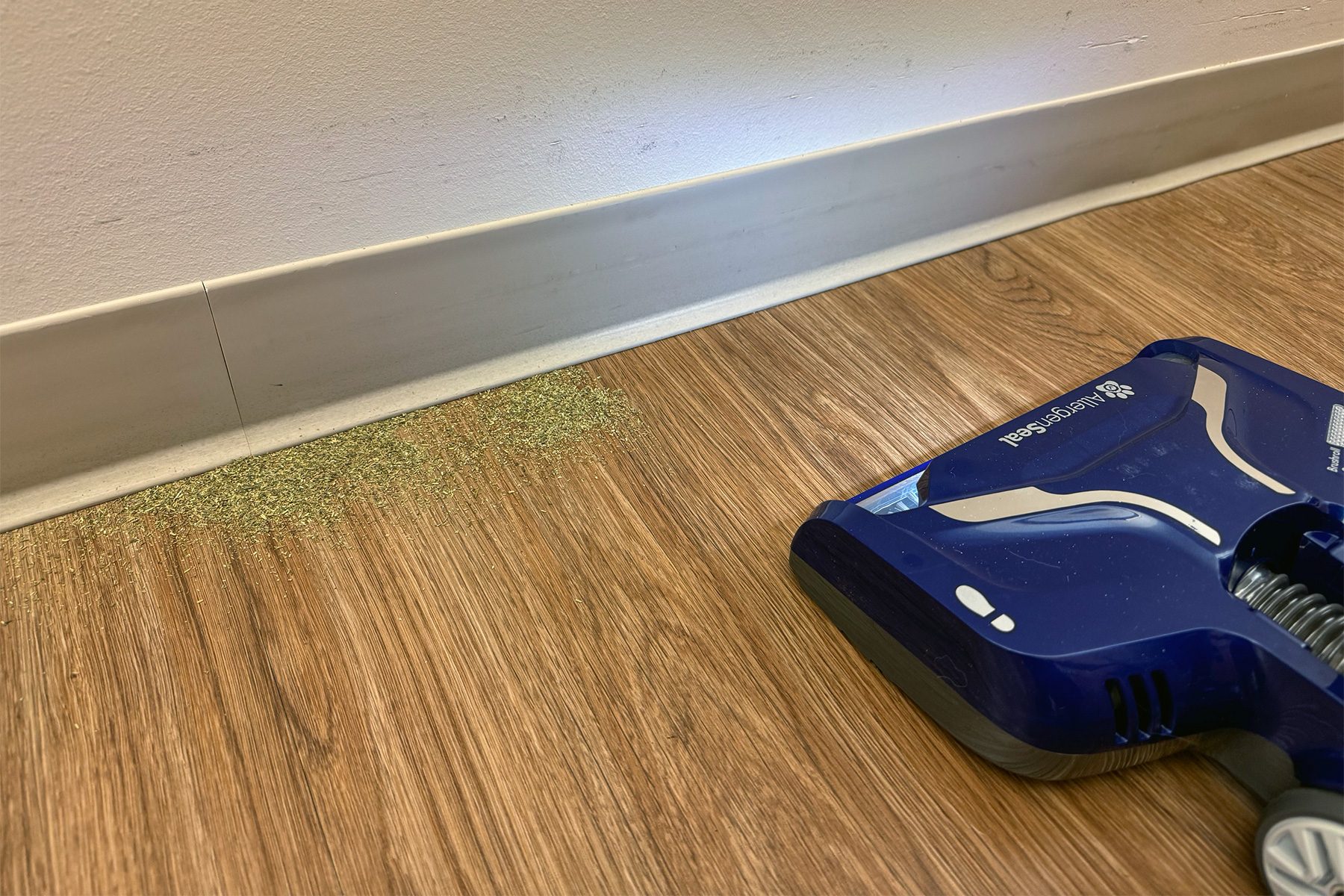Vacuum sweeper approaches scattered green herbs on wooden floor near a white wall.