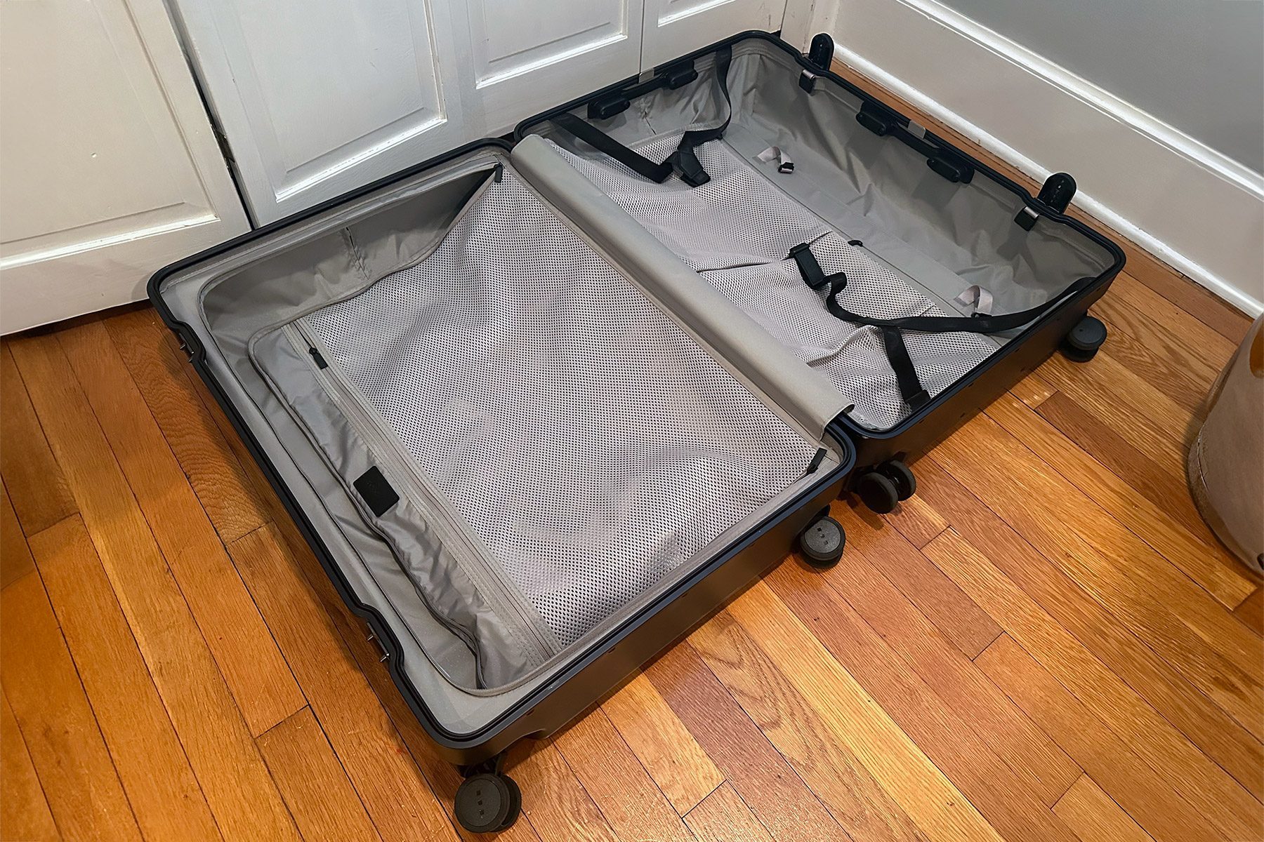 A suitcase lies open on a wooden floor, displaying empty compartments and straps inside, adjacent to white cupboard doors.