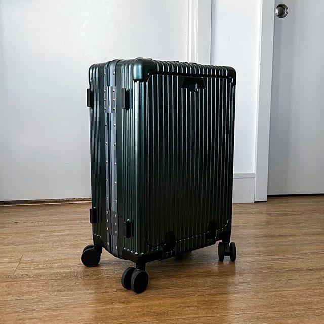 A dark green, hard-shell suitcase stands on wheels in a room with wooden flooring and a closed white door.