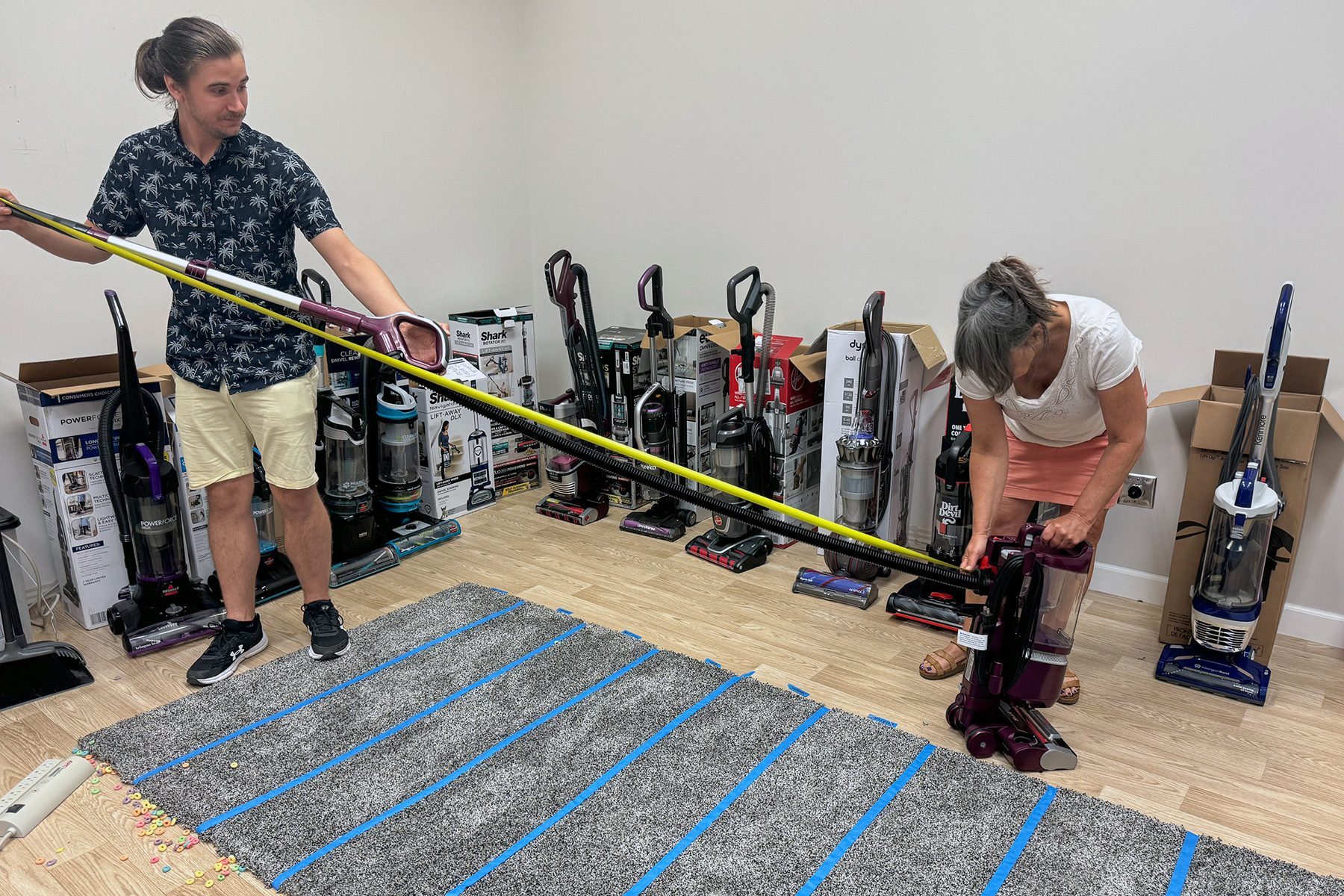 Vacuum cleaners sit in a row while two people measure the carpet using a tape measure on a hardwood floor.