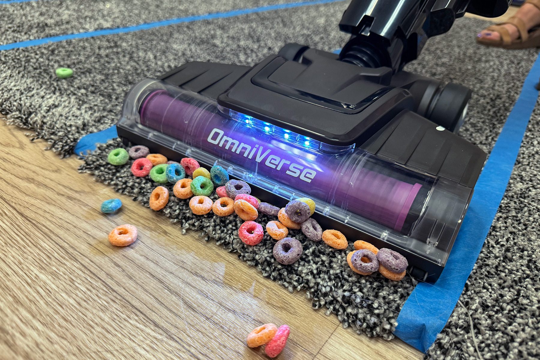 Vacuum cleaner picks up colorful cereal from a carpet's edge onto a wooden floor.