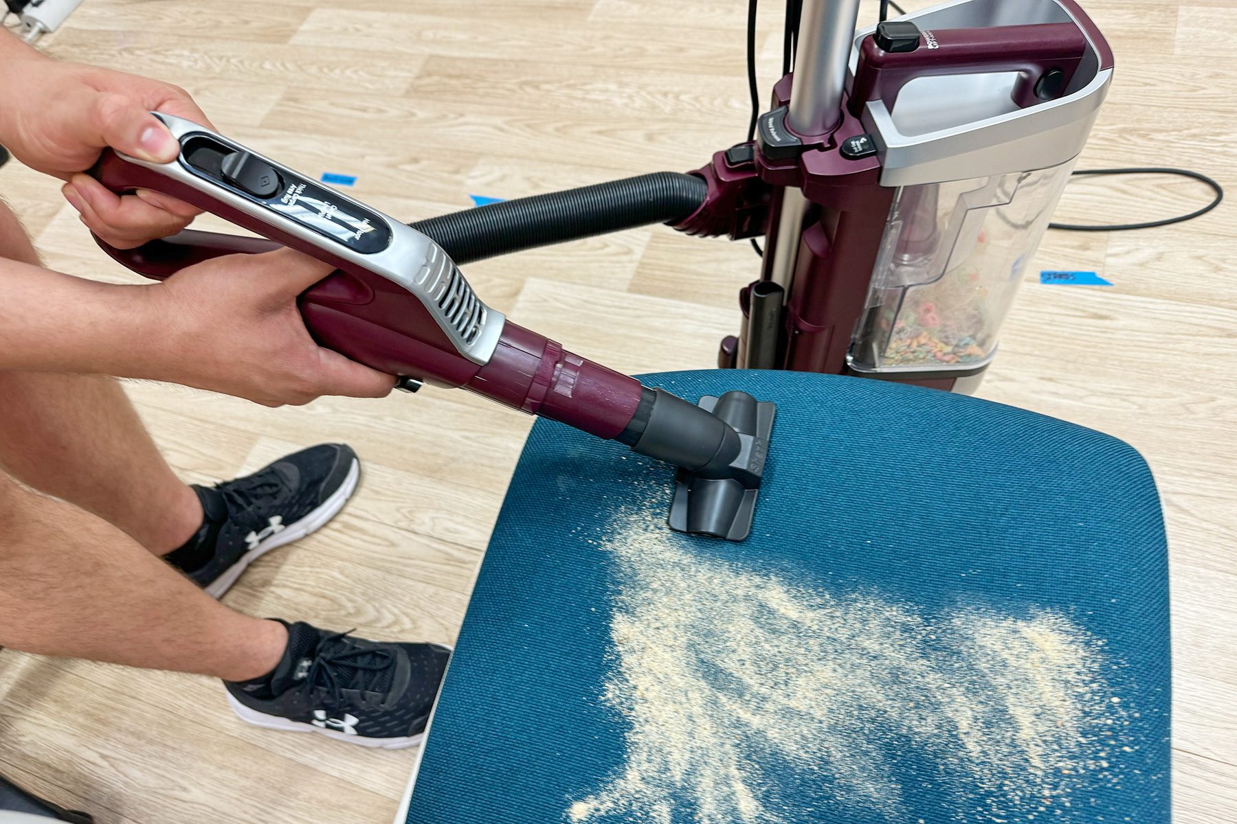 Vacuum removes debris from a blue cushioned chair, operated by hands, set in a room with light wood flooring.