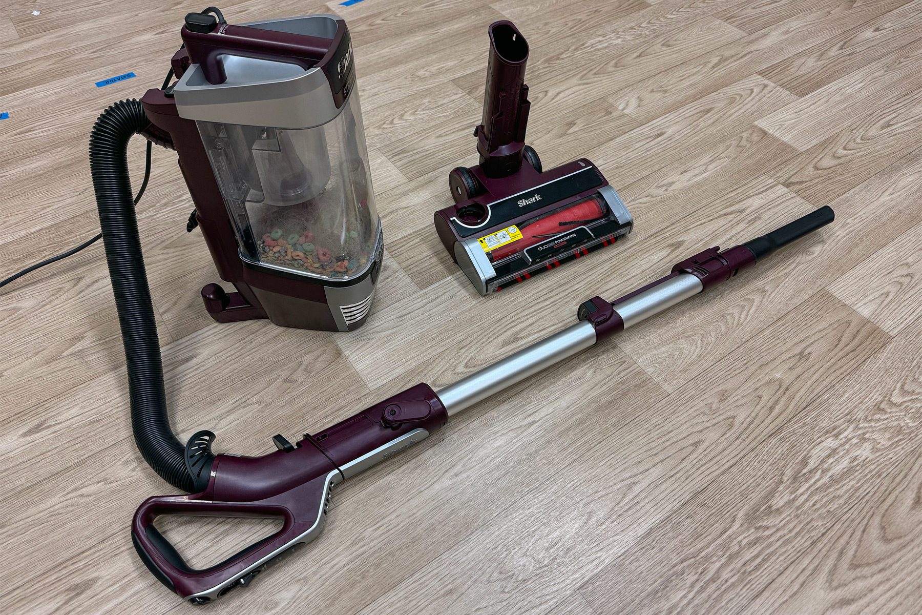 Vacuum components resting dismantled on wooden floor, showcasing a dirt-filled canister, flexible hose, and labeled base unit.