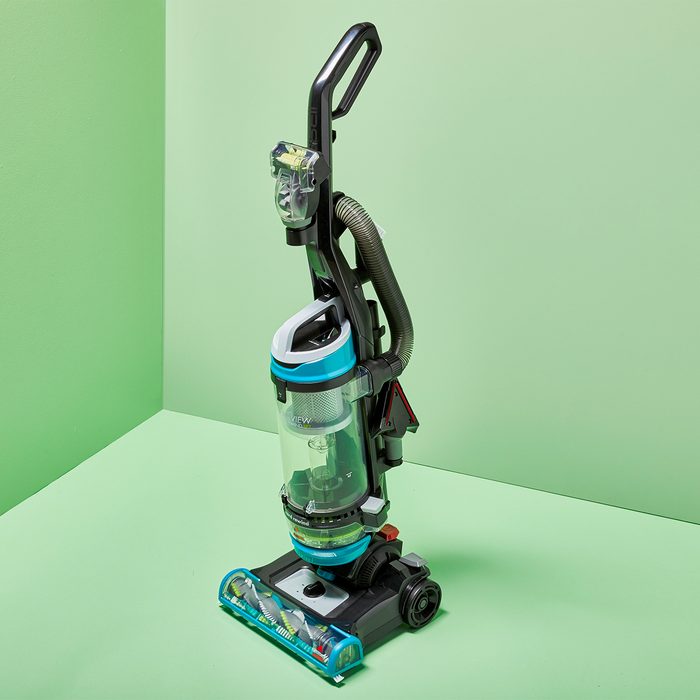 A blue and black upright vacuum cleaner stands on a light green cornered floor, with a visible handle and hose attachment.