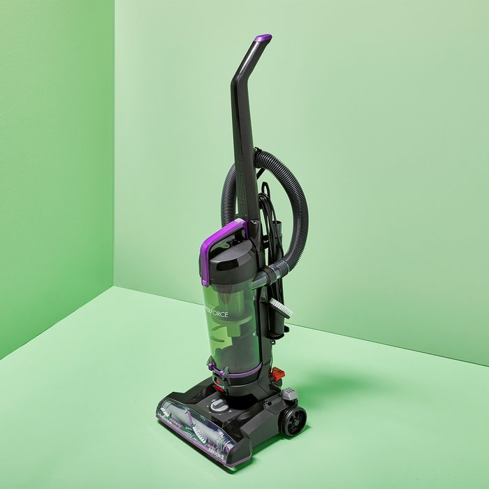 A vacuum cleaner stands upright in a corner with green walls, featuring purple accents and a transparent dust container.