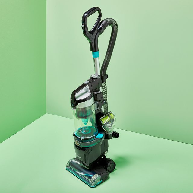 Vacuum cleaner stands upright in a green corner, featuring a clear canister and black handle.