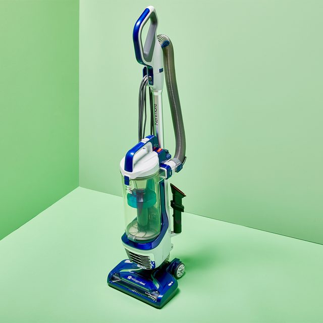 A vacuum cleaner stands upright on a green floor against a green wall, displaying a sleek, blue and silver design.