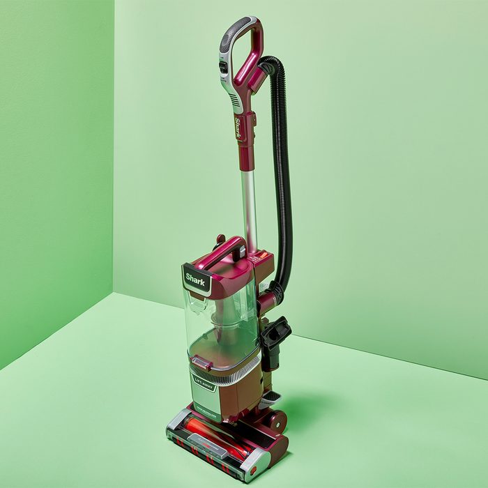 A red Shark vacuum cleaner stands upright against a light green wall, showcasing its sleek design and clear dust container.