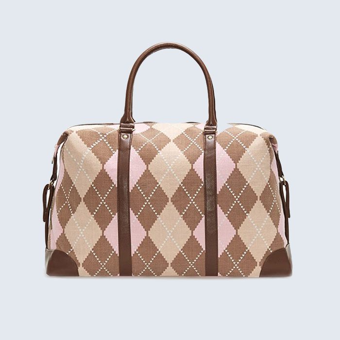 A patterned duffel bag rests on a light background, featuring brown and pink argyle design with leather handles and accents.