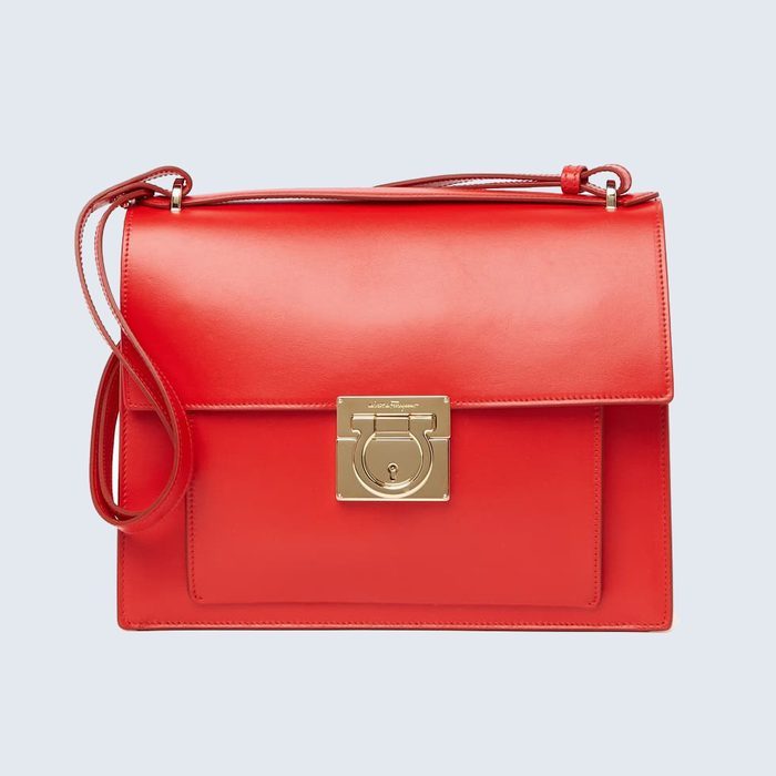 Red handbag displays gold clasp; strap drapes casually. Background is plain light blue.
