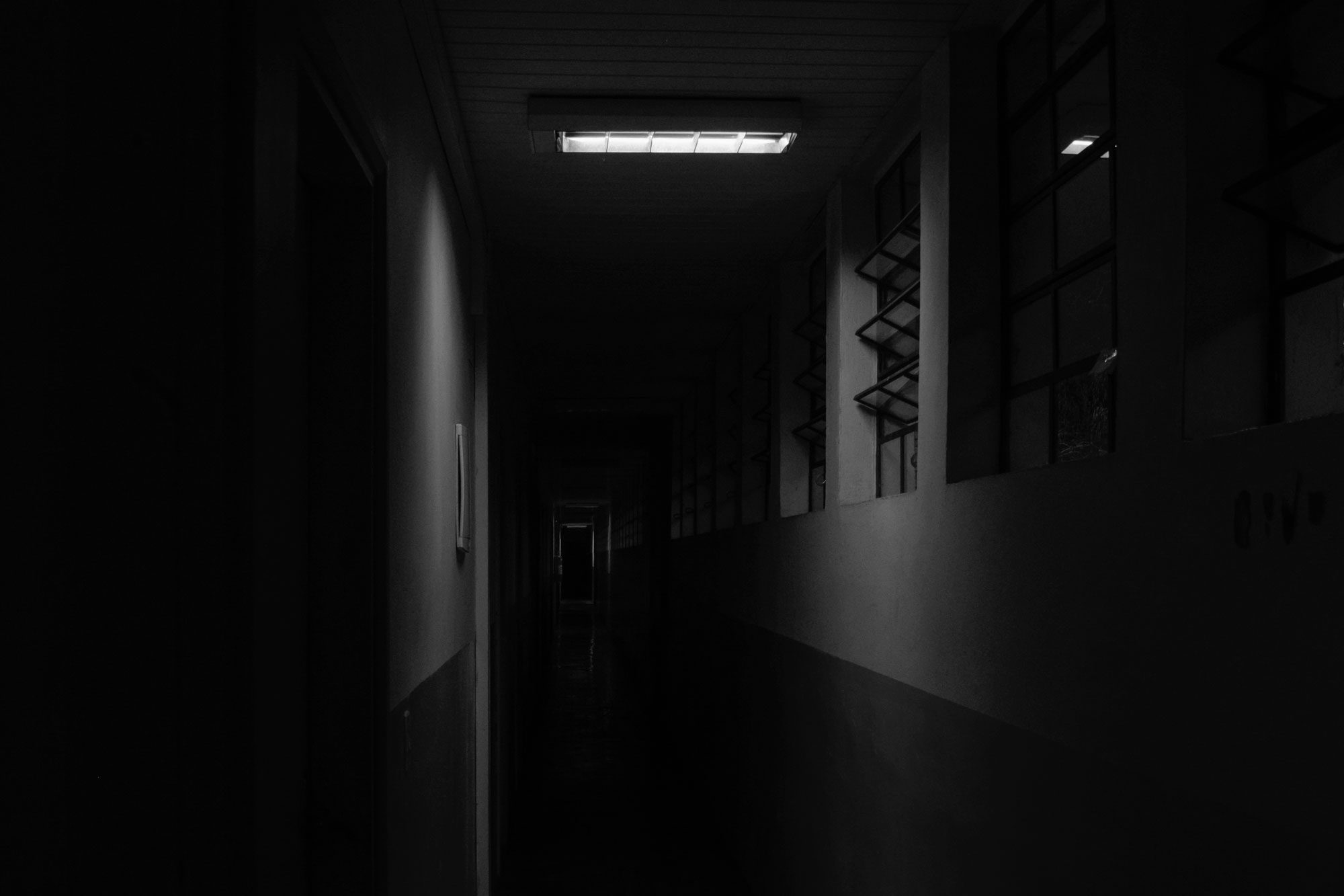 A dimly lit fluorescent tube illuminates a narrow, dark corridor with barred windows along one side.